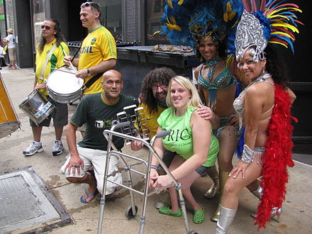 A seven people are seen at Chicago's annual Disability Pride Parade in 2011. In the center, a woman with a visible physical disability sits next to a walker while other attendees in costume hold instruments around her.